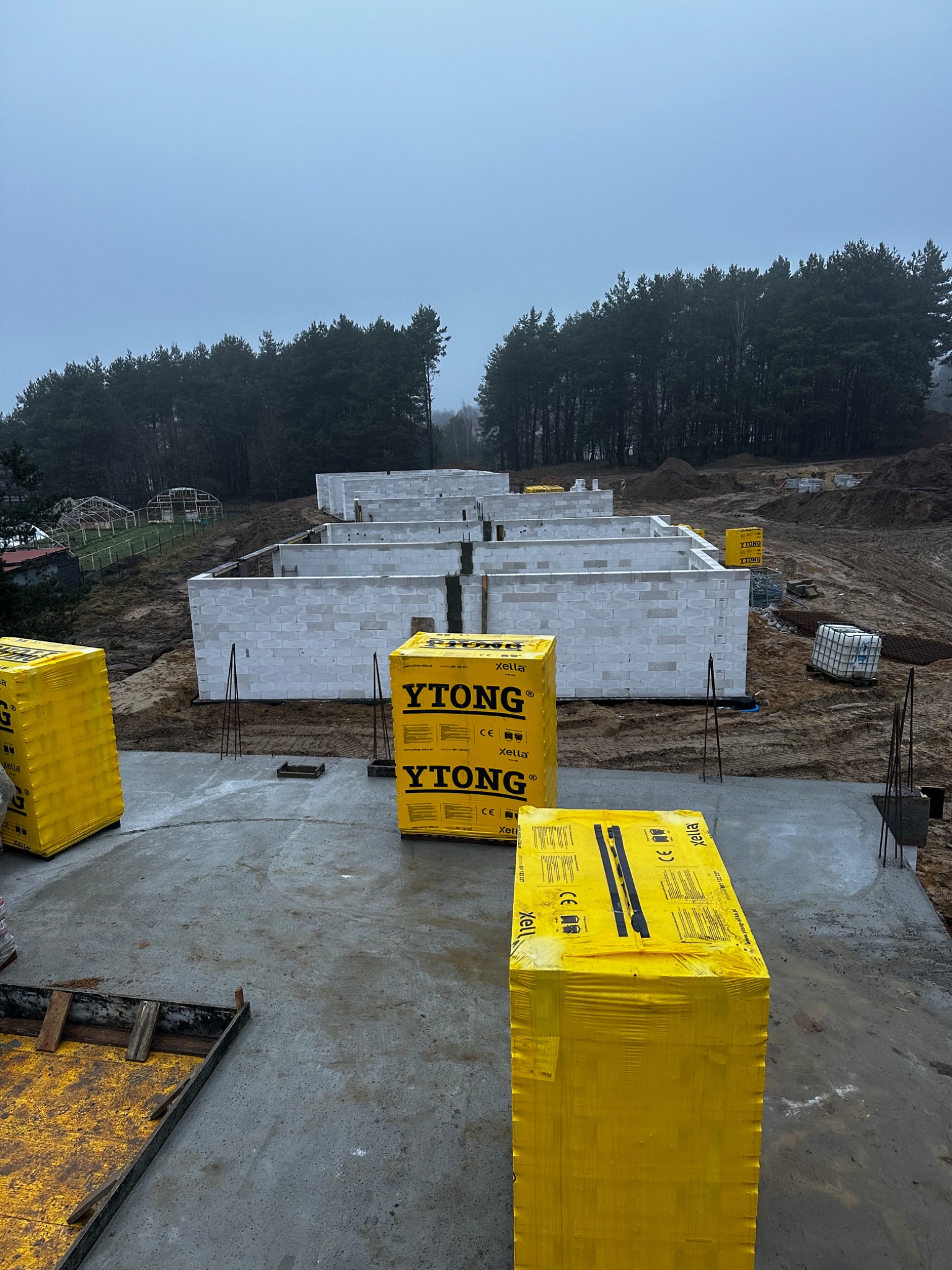 Fundamenty domu z betonu komórkowego Ytong, widoczne żółte palety z bloczkami. W tle las i pochmurne niebo. Etap budowy domu.