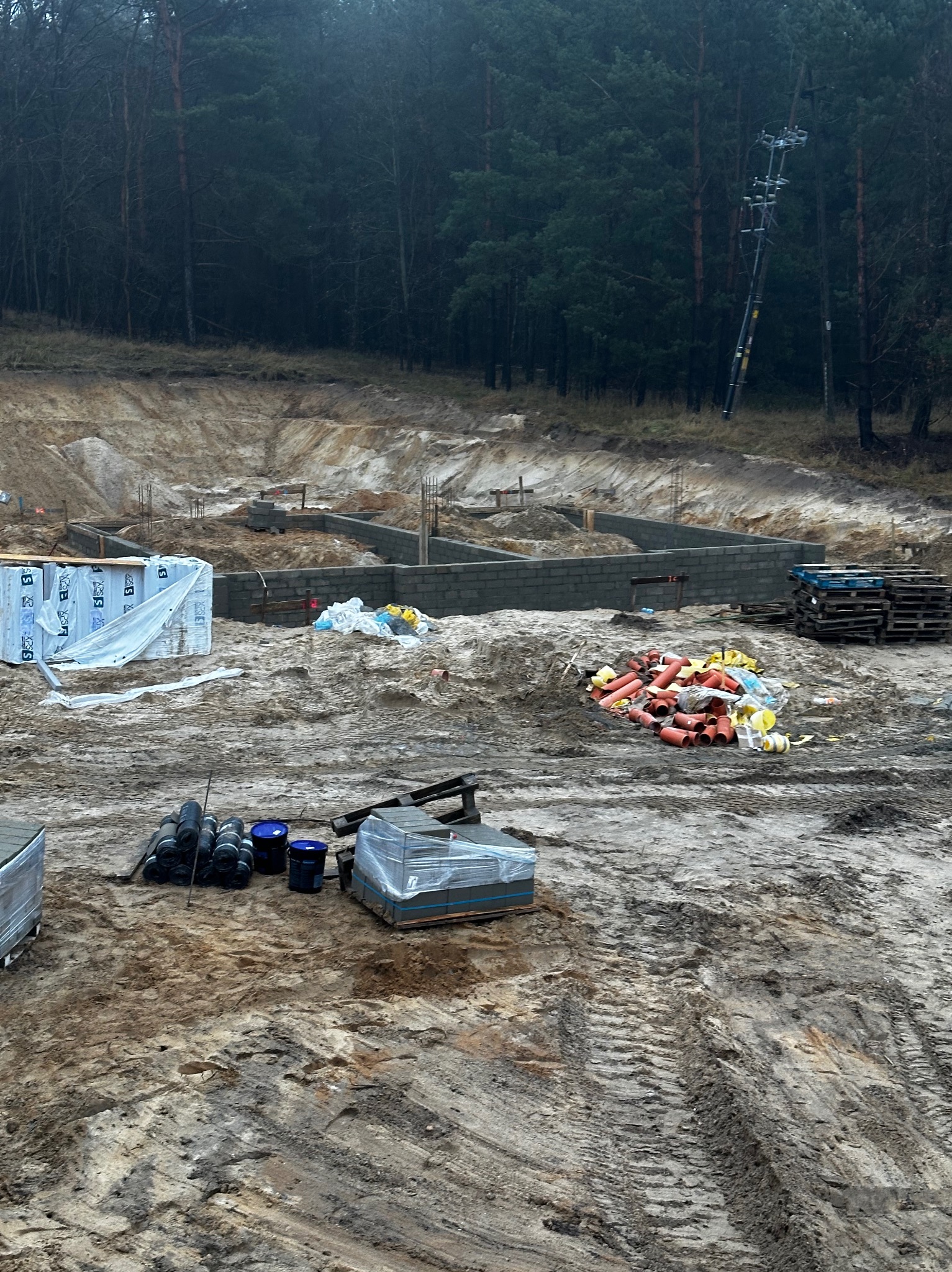 Fundamenty domu z bloczków betonowych na tle lasu, widoczne materiały budowlane i wykop. Etap budowy w trakcie realizacji, teren przygotowany pod dalsze prace.