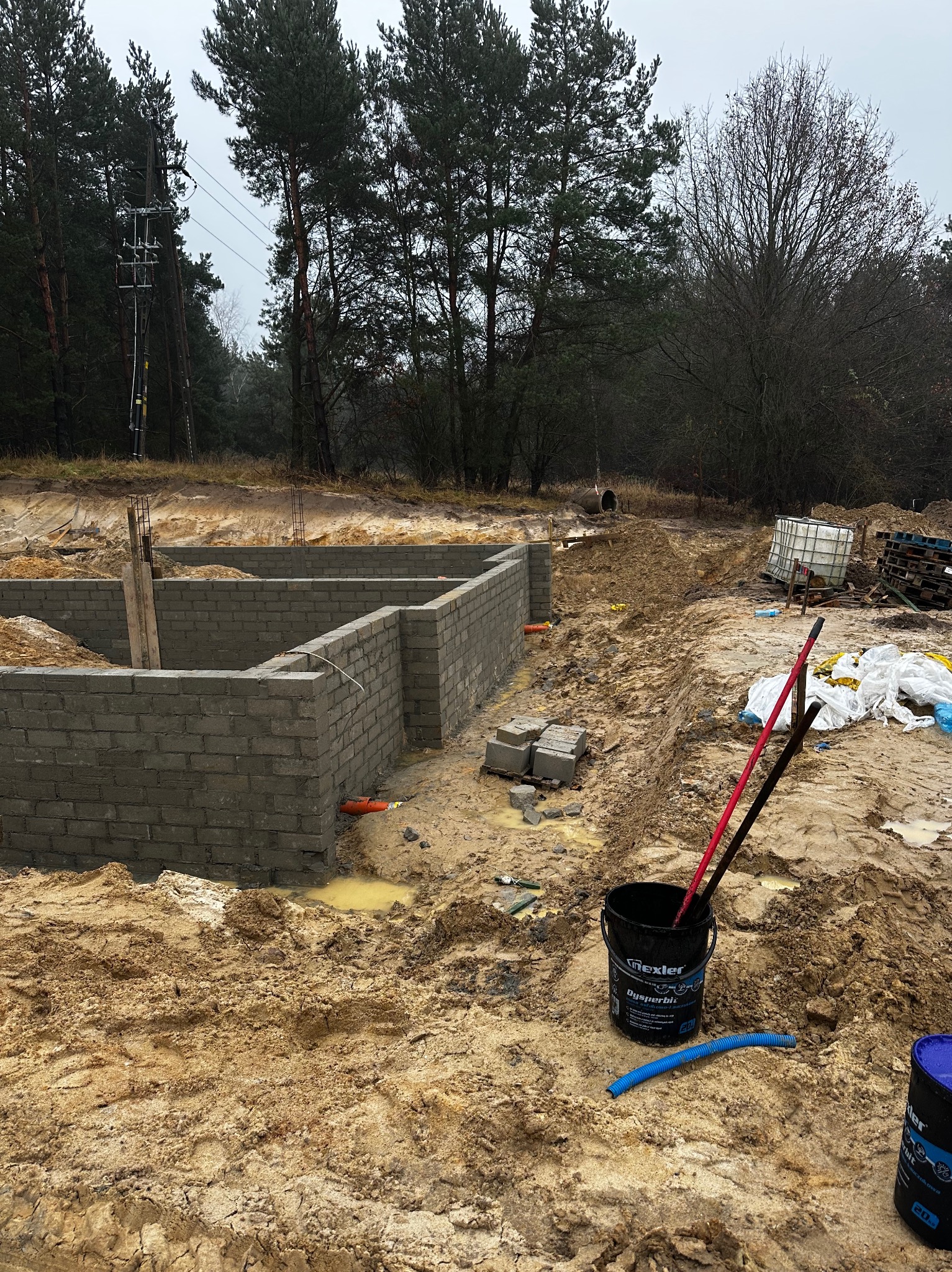 Wykop pod fundamenty z częściowo wymurowanymi ścianami z szarego bloczka betonowego. Widoczne narzędzia i materiały budowlane. Teren budowy w lesie.