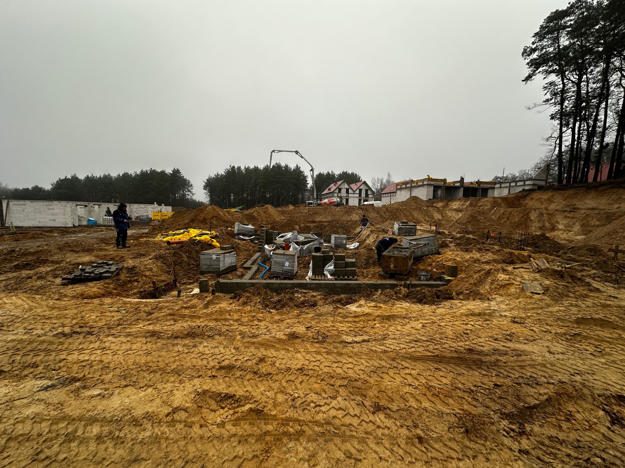 Fundamenty domu w trakcie budowy, widoczne bloczki i betonowa pompa na tle lasu i pochmurnego nieba. Prace murarskie w toku, teren budowy z wykopami i materiałami.