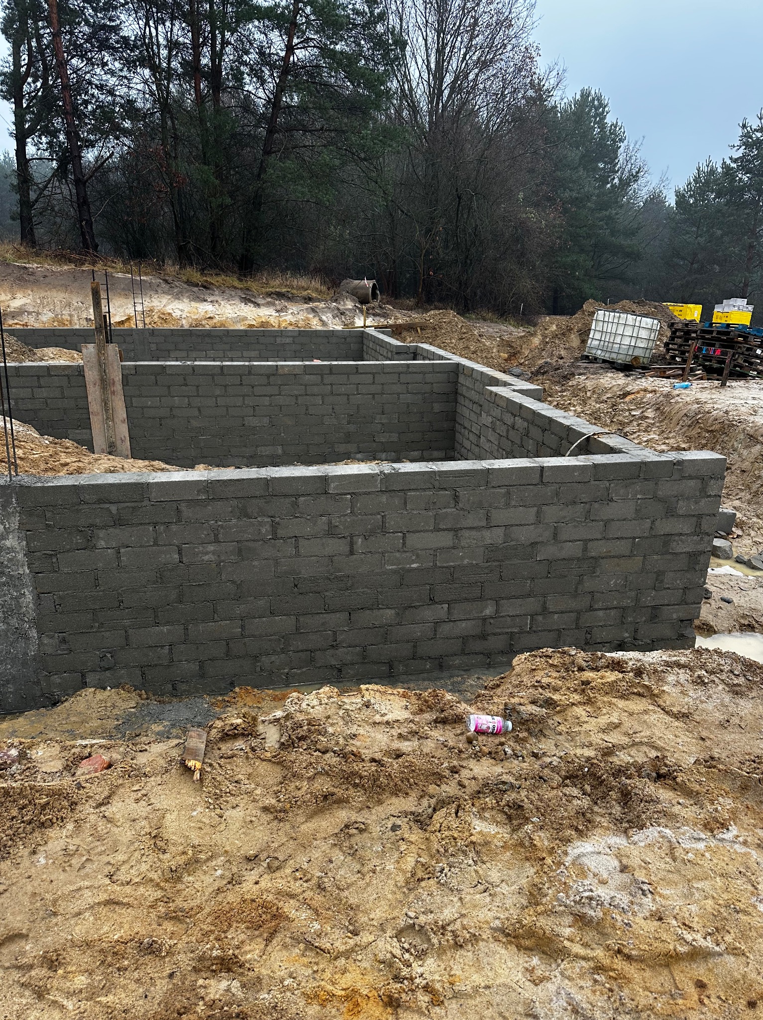 Wzniesione mury fundamentowe z szarego bloczka betonowego na zewnątrz, w tle drzewa i palety z pojemnikami. Widok z poziomu gruntu.