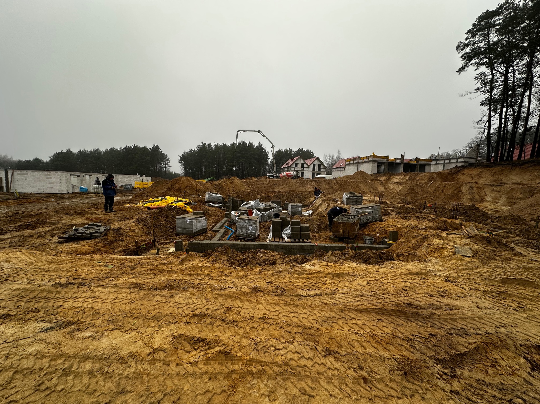 Fundamenty domu w trakcie budowy, widoczne bloczki fundamentowe, ziemia i sprzęt budowlany. W tle las i częściowo wybudowane ściany.