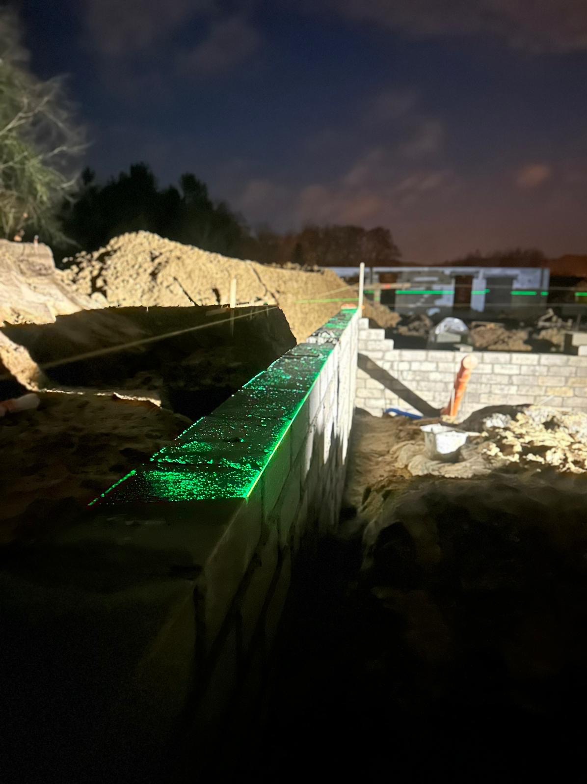 Nocne murowanie: Świecąca zielona linia lasera wyznacza poziom na wznoszonej ścianie z bloczków, w tle hałda piasku i zarys budynku w trakcie budowy.