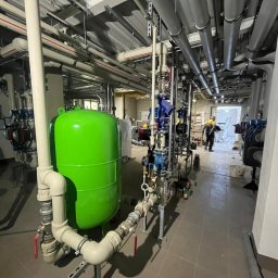 DAS PRO SPÓŁKA Z OGRANICZONĄ ODPOWIEDZIALNOŚCIĄ - Kotłownia z zielonym zbiornikiem ciśnieniowym, licznymi rurami i zaworami. W tle pracownik w żółtym ubraniu. Szara podłoga i sufit z instalacjami.