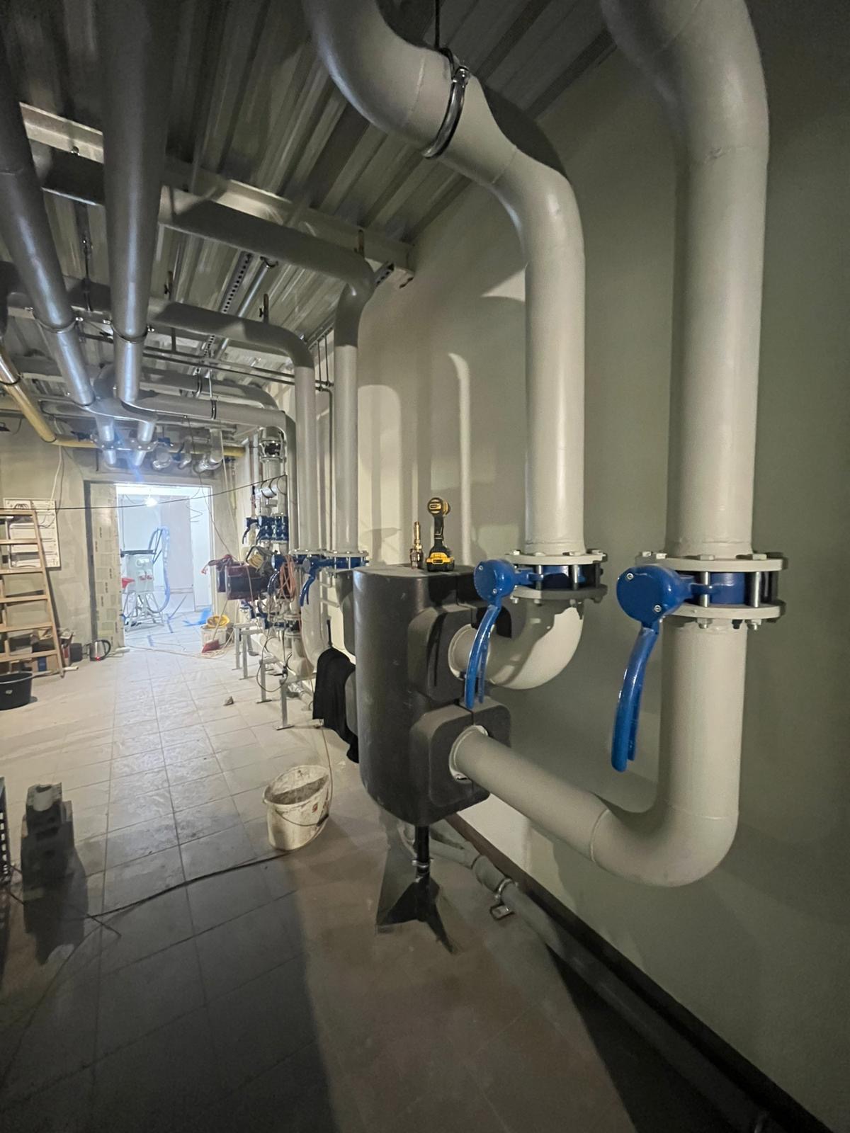 Instalacja przemysłowa z rurami, zaworami z niebieskimi uchwytami i narzędziem akumulatorowym na filtrze. Widoczna drabina i sprzęt w tle.