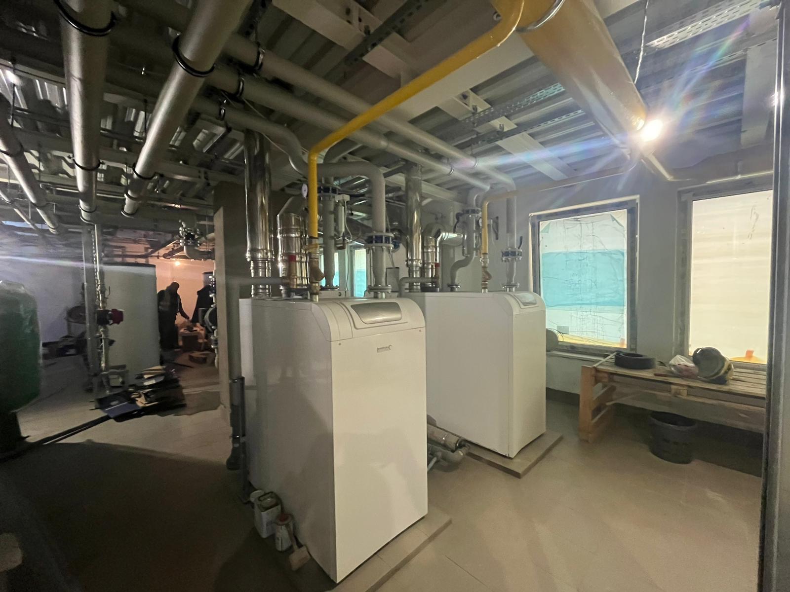 Dwa białe piece CO z żółtymi rurami gazowymi w pomieszczeniu technicznym. Widoczne rury, okna zakryte folią i pracownicy w tle.