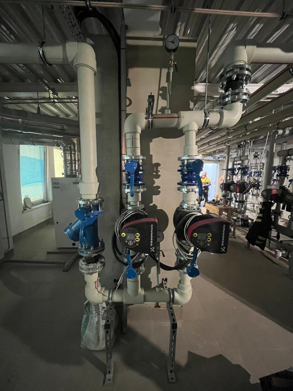 Złożona instalacja hydrauliczna z dwoma pompami Grundfos, zaworami i rurami z tworzywa, widoczny manometr i elementy montażowe. W tle instalator przy oknie.