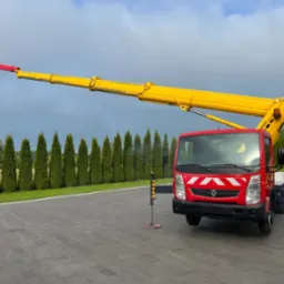 Czerwony podnośnik koszowy z żółtym ramieniem na tle zielonych drzew i szarej kostki brukowej. Ramię podniesione, kosz na końcu ramienia. Stabilizatory rozłożone.