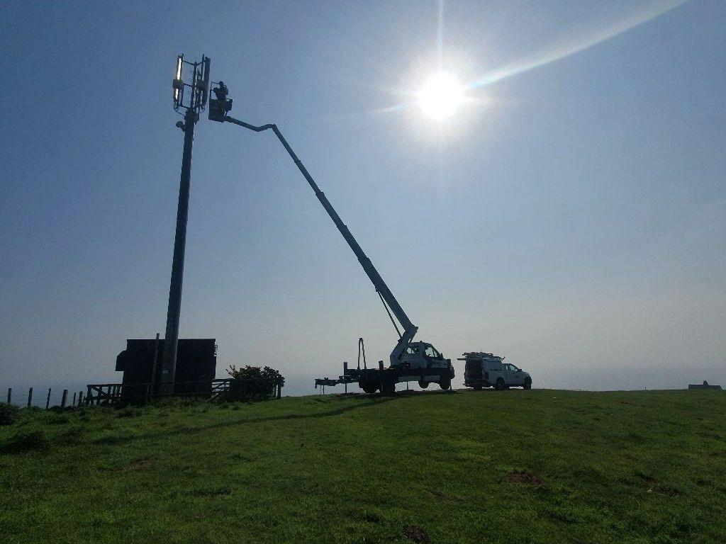 Praca na wysokości: Podnośnik koszowy z pracownikiem przy maszcie telekomunikacyjnym na wzgórzu, z widokiem na morze. Słoneczny dzień.