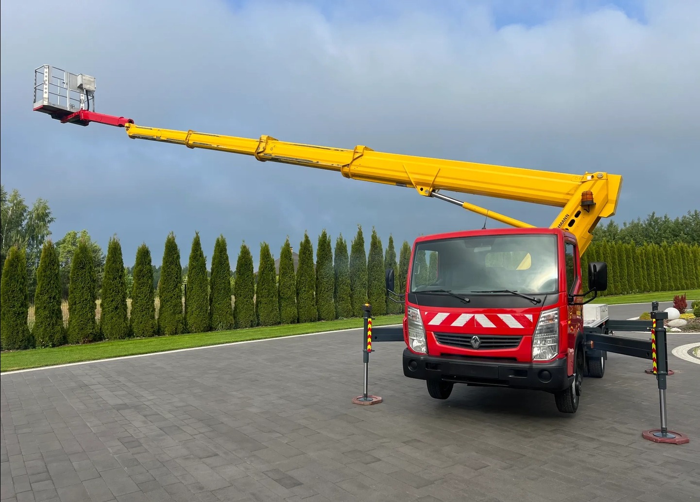 Czerwony podnośnik koszowy z żółtym ramieniem na tle zielonych drzew i szarej kostki brukowej. Ramię podniesione, kosz na końcu ramienia. Stabilizatory rozłożone.