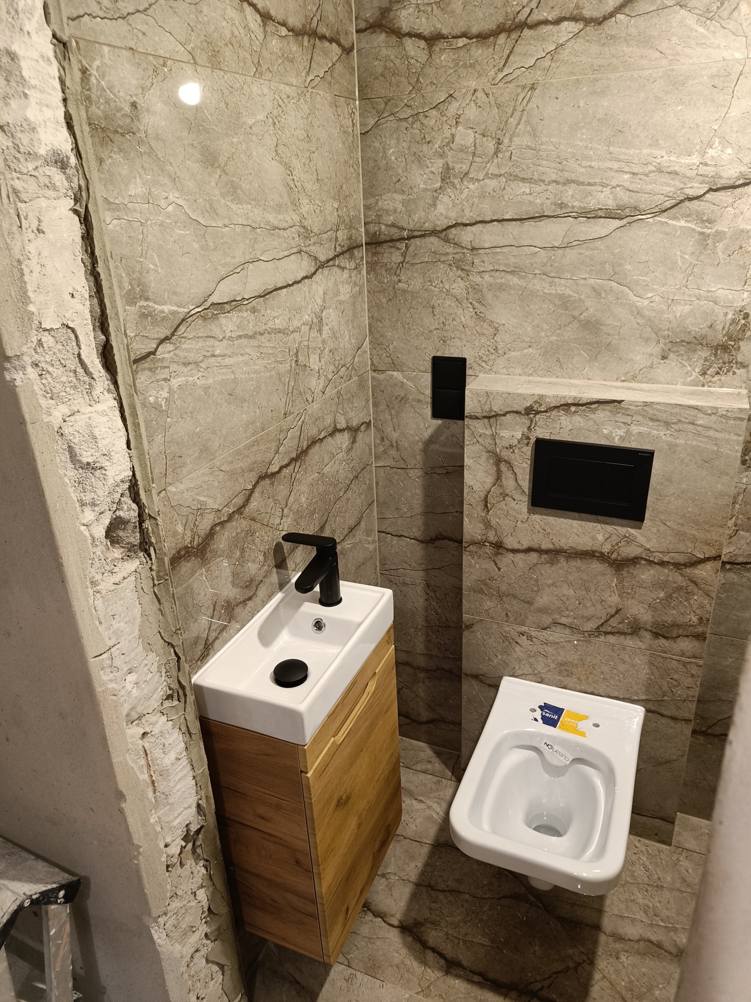 Łazienka w trakcie wykończenia: marmurowe płytki, wisząca toaleta, mała umywalka z drewnianą szafką i czarną armaturą. Ściana obok umywalki w stanie surowym.