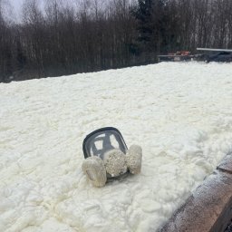 Arkadiusz Adacha ADACHA tynki i izolacje - Poddasze po ociepleniu pianką PUR, widoczna faktura piany i pozostawiona maska ochronna. W tle drzewa i elementy konstrukcyjne dachu.