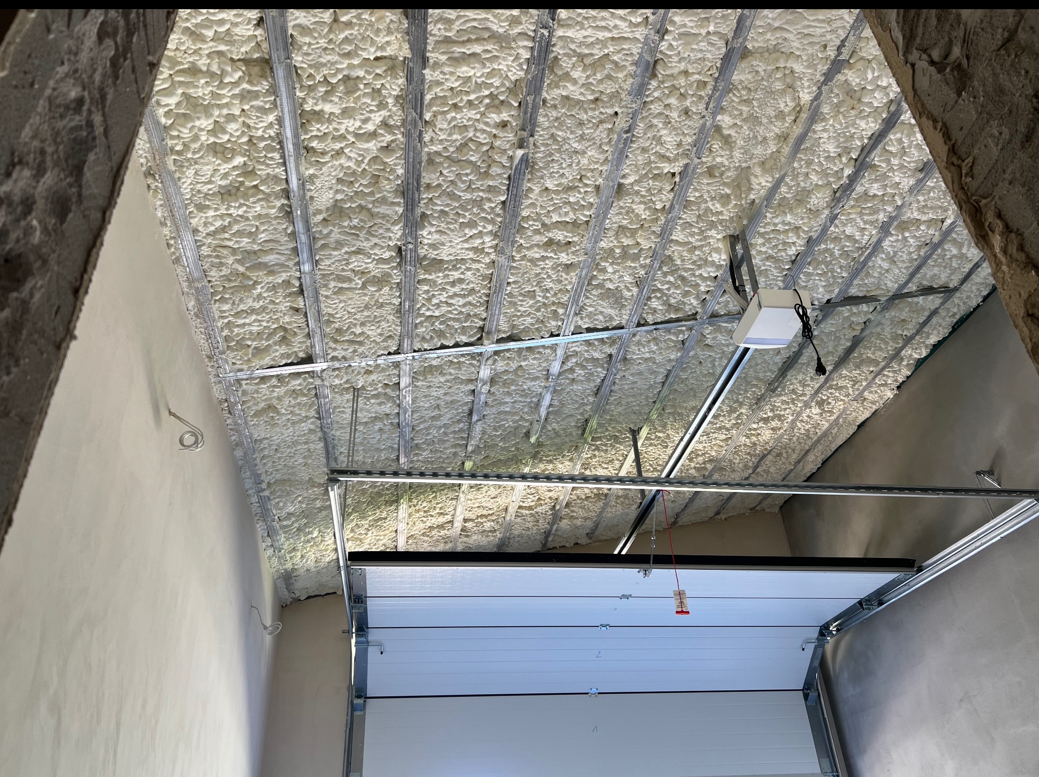 Poddasze z widoczną izolacją pianką PUR między metalowymi profilami, częściowo opuszczona brama garażowa. Ujęcie z dołu, widoczne fragmenty ścian.
