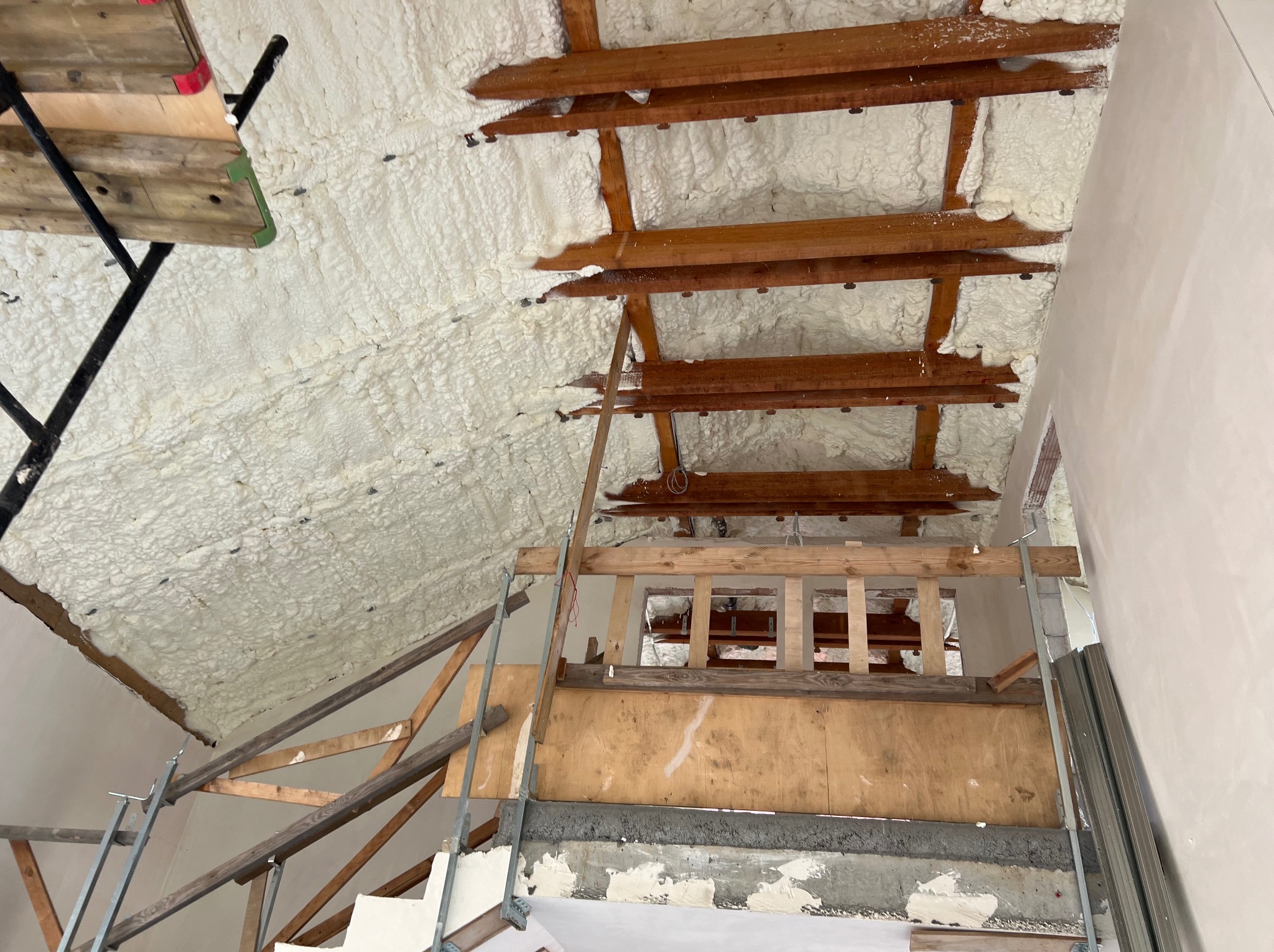 Poddasze z drewnianą konstrukcją pokryte białą pianką izolacyjną, widoczne rusztowanie. Izolacja wykonana na nierównej powierzchni.
