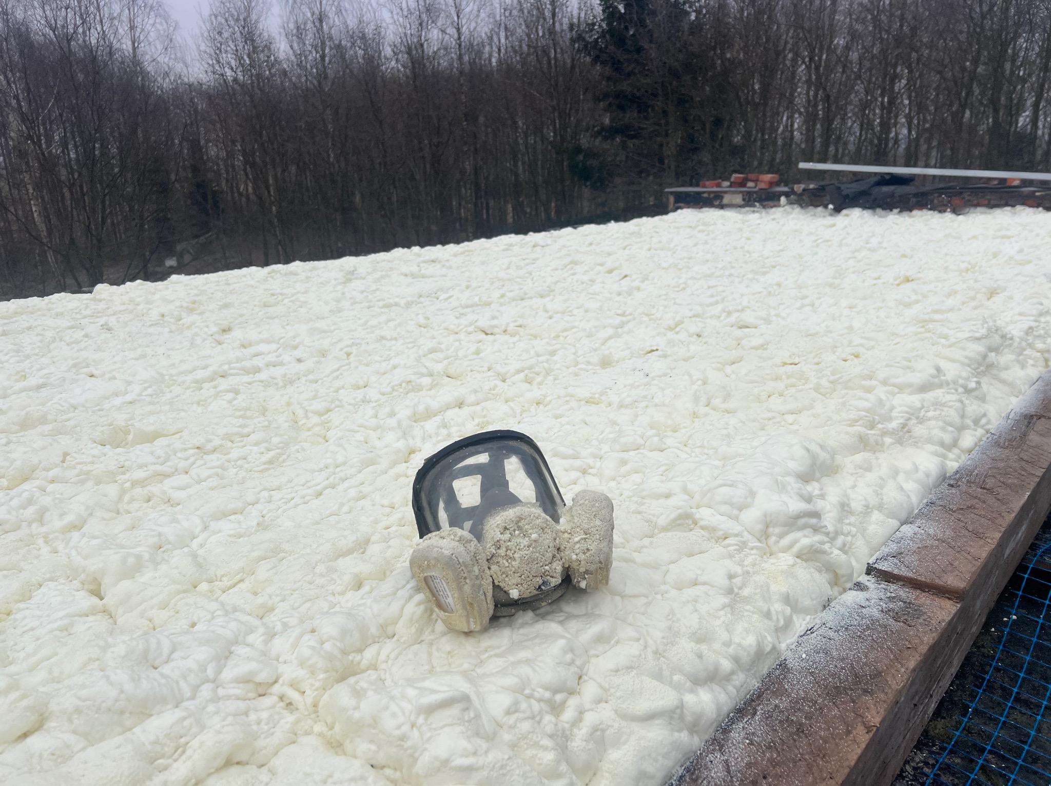Poddasze po ociepleniu pianką PUR, widoczna faktura piany i pozostawiona maska ochronna. W tle drzewa i elementy konstrukcyjne dachu.