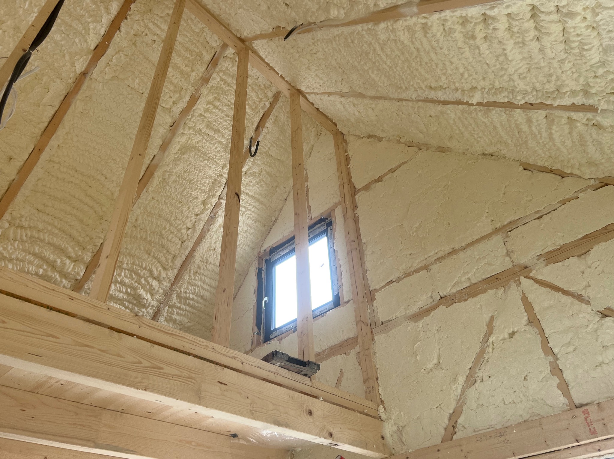 Poddasze ocieplone pianką PUR, widoczne drewniane elementy konstrukcyjne i okno dachowe. Biała, nierówna struktura pianki wypełnia przestrzenie między belkami.