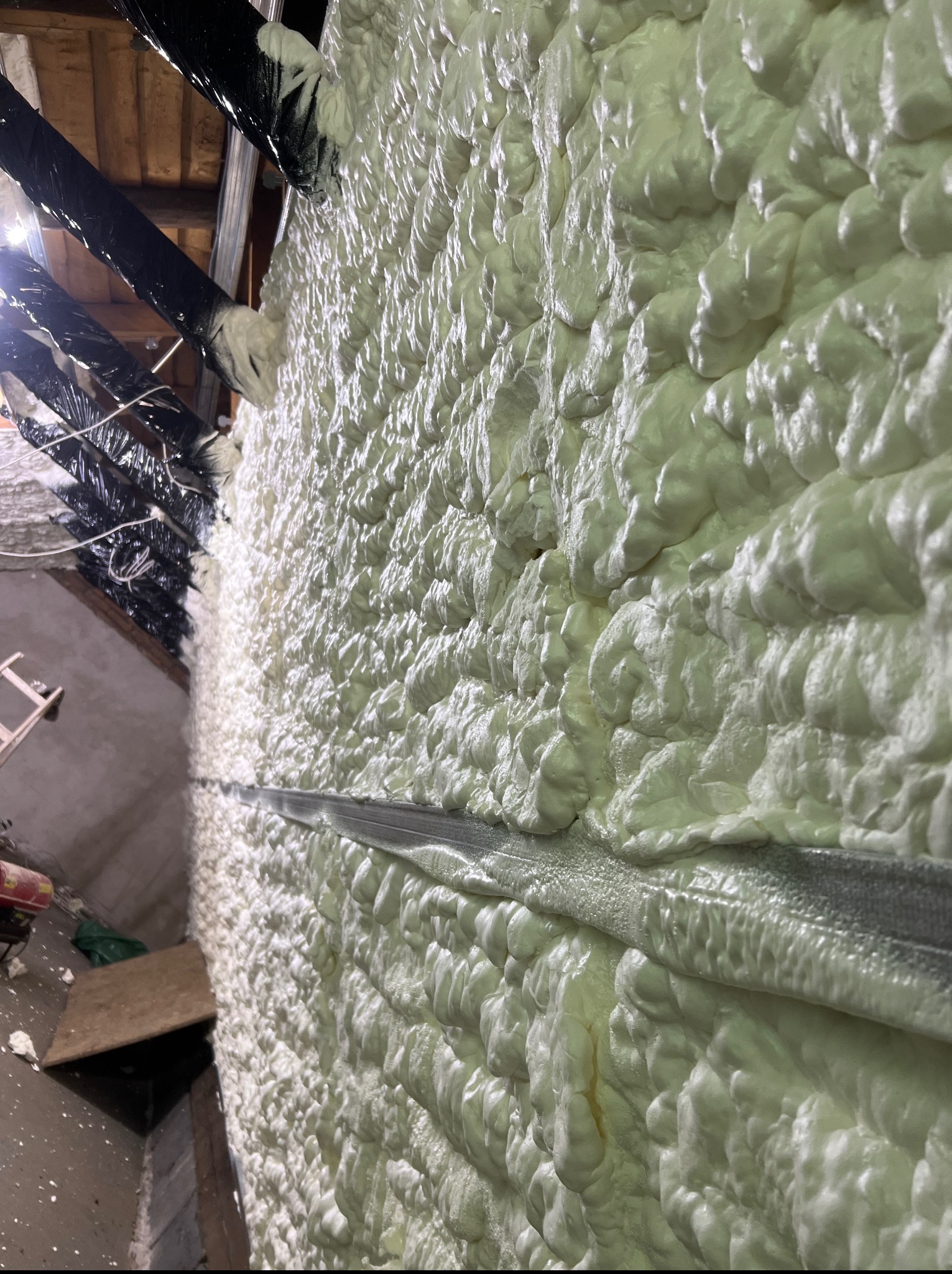 Świeżo nałożona, jasna pianka poliuretanowa szczelnie wypełnia przestrzeń poddasza, przylegając do metalowego profilu. Widoczne elementy konstrukcji dachu i instalacji.