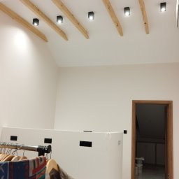 REM-NOV Michał Nowacki - Jasne wnętrze z drewnianymi belkami stropowymi i nowoczesnym oświetleniem. Widoczna biała ściana, marmurowe płytki podłogowe i fragment garderoby.