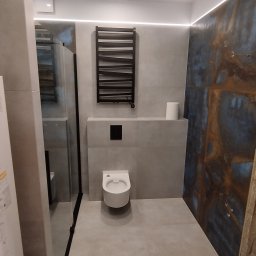 REM-NOV Michał Nowacki - Nowoczesna łazienka z szarymi płytkami, podwieszaną toaletą i grzejnikiem drabinkowym. Dekoracyjna ściana z efektem rdzy. Oświetlenie LED podkreśla minimalistyczny design.