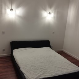 REM-NOV Michał Nowacki - Nowoczesna sypialnia z drewnianą podłogą, czarnym łóżkiem i białymi ścianami oświetlonymi przez dwa kinkiety. W tle widoczny kryształowy żyrandol.