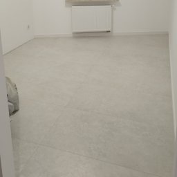 REM-NOV Michał Nowacki - Jasne, matowe płytki podłogowe w nowym, minimalistycznym wnętrzu. Widoczny grzejnik i białe listwy przypodłogowe. W rogu pomieszczenia worek z materiałami.