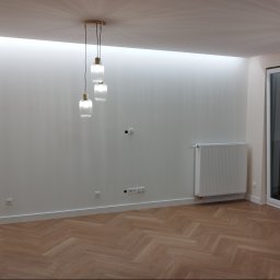 REM-NOV Michał Nowacki - Jasne wnętrze po remoncie: parkiet w jodełkę, białe ściany z gładzią, nowoczesne oświetlenie sufitowe i grzejnik. Widoczne gniazdka elektryczne i listwy przypodłogowe.
