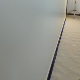 REM-NOV Michał Nowacki - Gładka, świeżo pomalowana ściana w jasnym odcieniu szarości, zabezpieczona niebieską taśmą malarską przy podłodze. W tle widoczny statyw oświetleniowy.