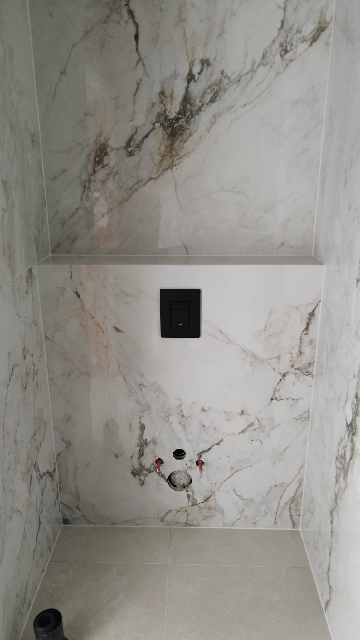 Wykończona toaleta w łazience z marmurowymi płytkami, czarnym przyciskiem spłukującym i przygotowanymi przyłączami wodno-kanalizacyjnymi.