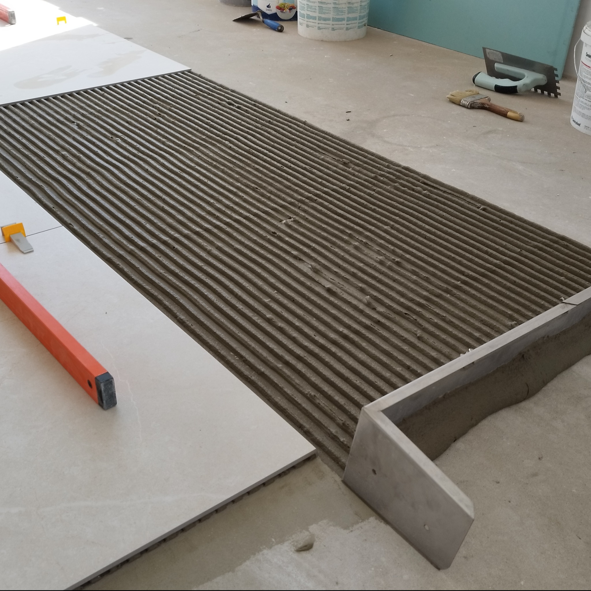 Przygotowanie podłoża pod płytki: równomiernie rozprowadzony klej na podłodze, częściowo ułożone płytki, poziomica i narzędzia glazurnicze w tle.