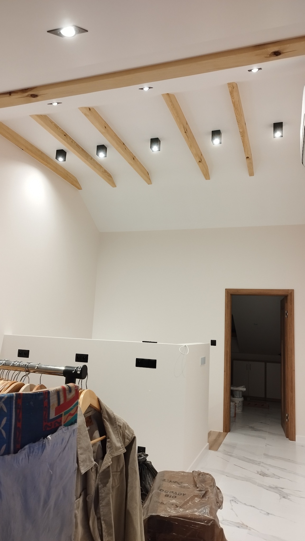 Jasne wnętrze z drewnianymi belkami stropowymi i nowoczesnym oświetleniem. Widoczna biała ściana, marmurowe płytki podłogowe i fragment garderoby.