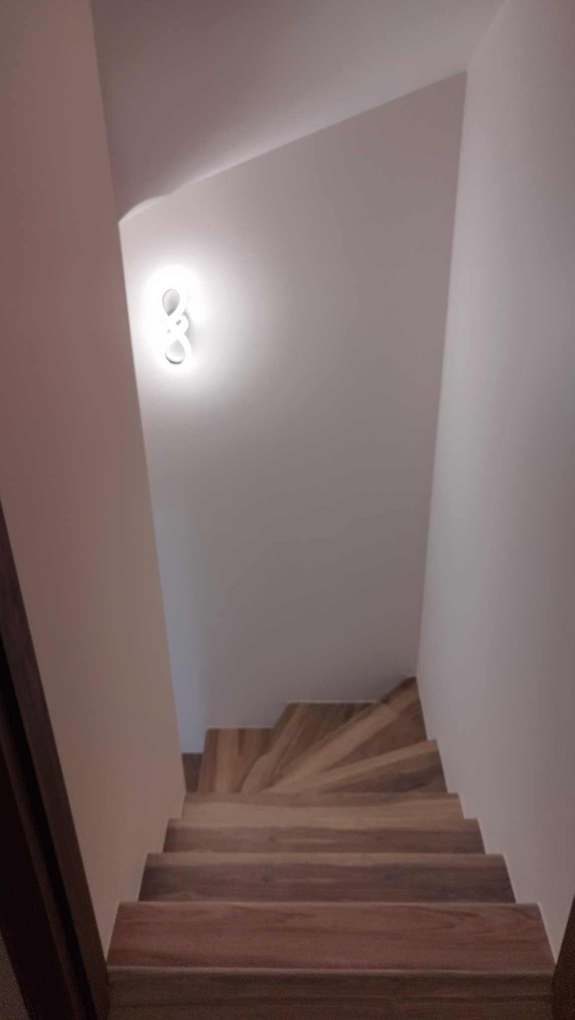 Drewniane schody w jasnym wnętrzu, oświetlone lampą sufitową. Widok z dołu, ukazujący zakręt schodów i minimalistyczne wykończenie ścian.