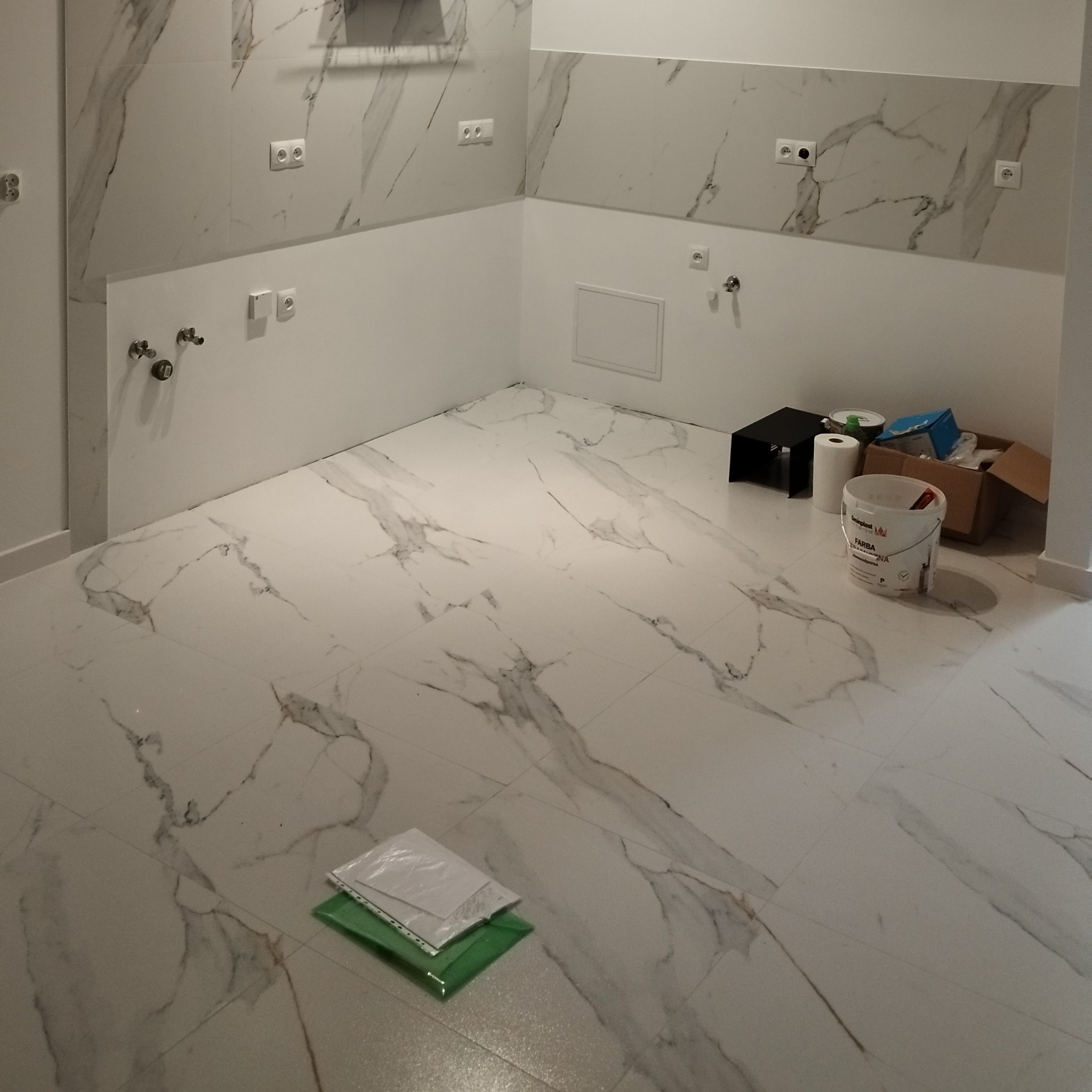 Wykończona łazienka z marmurowymi płytkami na ścianach i podłodze. Widoczne puszki elektryczne, instalacja wodna i akcesoria malarskie. Nowoczesny design.