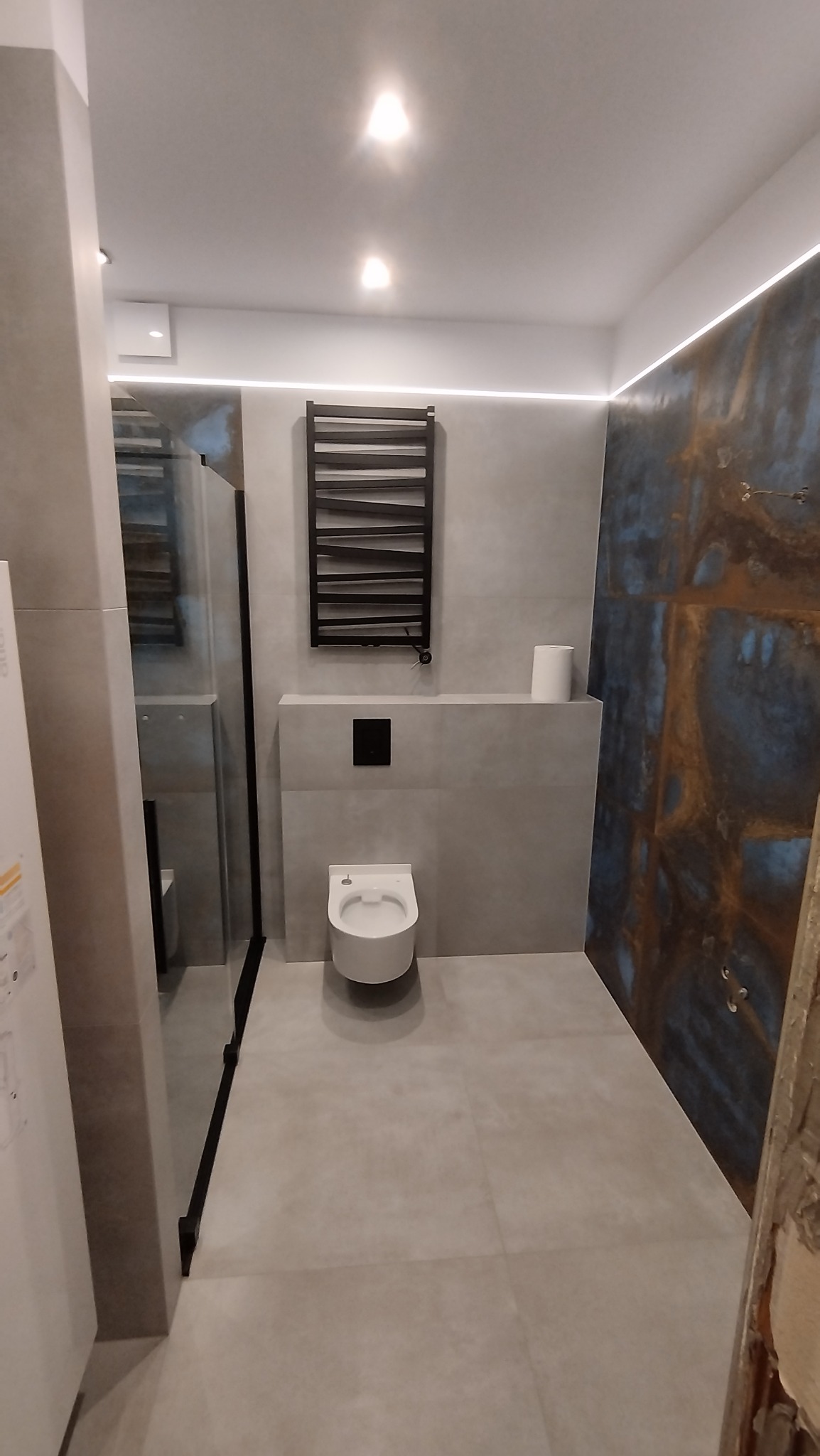 Nowoczesna łazienka z szarymi płytkami, podwieszaną toaletą i grzejnikiem drabinkowym. Dekoracyjna ściana z efektem rdzy. Oświetlenie LED podkreśla minimalistyczny design.