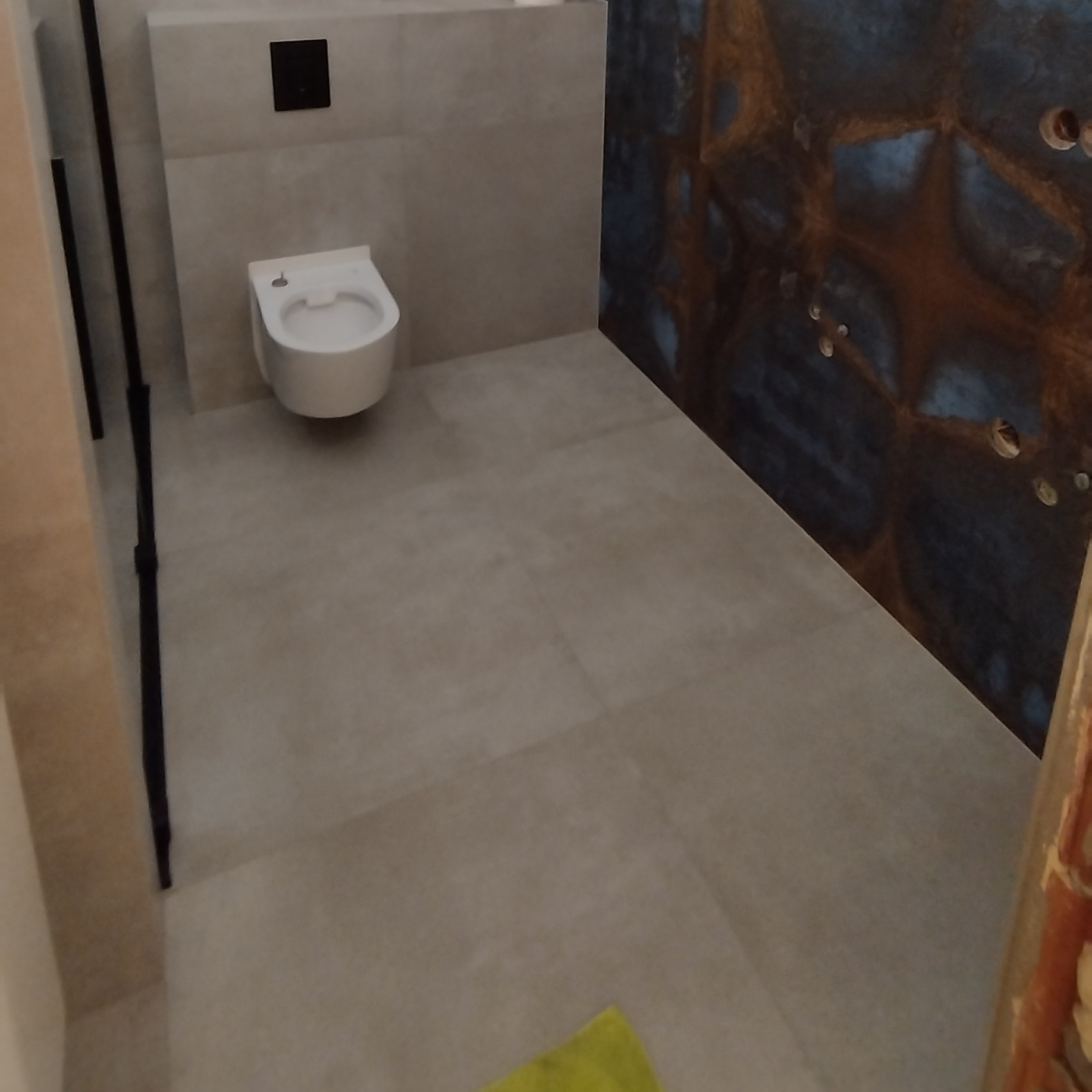 Nowoczesna łazienka z podwieszaną toaletą, beżowymi płytkami i dekoracyjną ścianą z efektem rdzy. Minimalistyczny design, widoczny fragment zielonego dywanika.