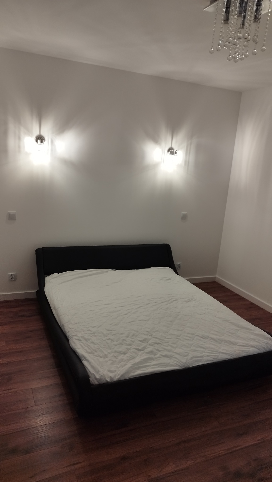 Nowoczesna sypialnia z drewnianą podłogą, czarnym łóżkiem i białymi ścianami oświetlonymi przez dwa kinkiety. W tle widoczny kryształowy żyrandol.