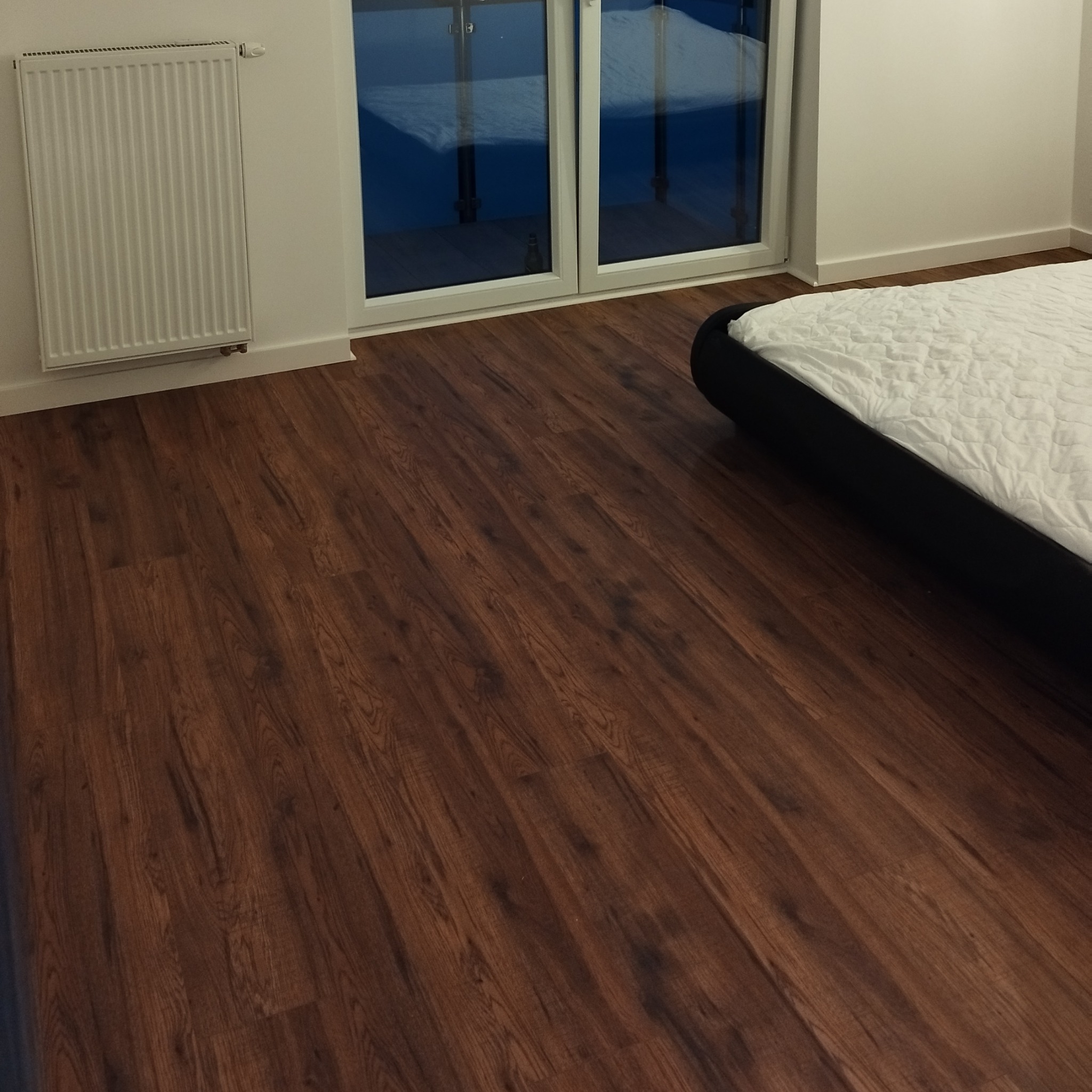 Ciemnobrązowe panele podłogowe ułożone w pokoju z białymi ścianami, grzejnikiem i fragmentem łóżka. Widoczne drzwi balkonowe.