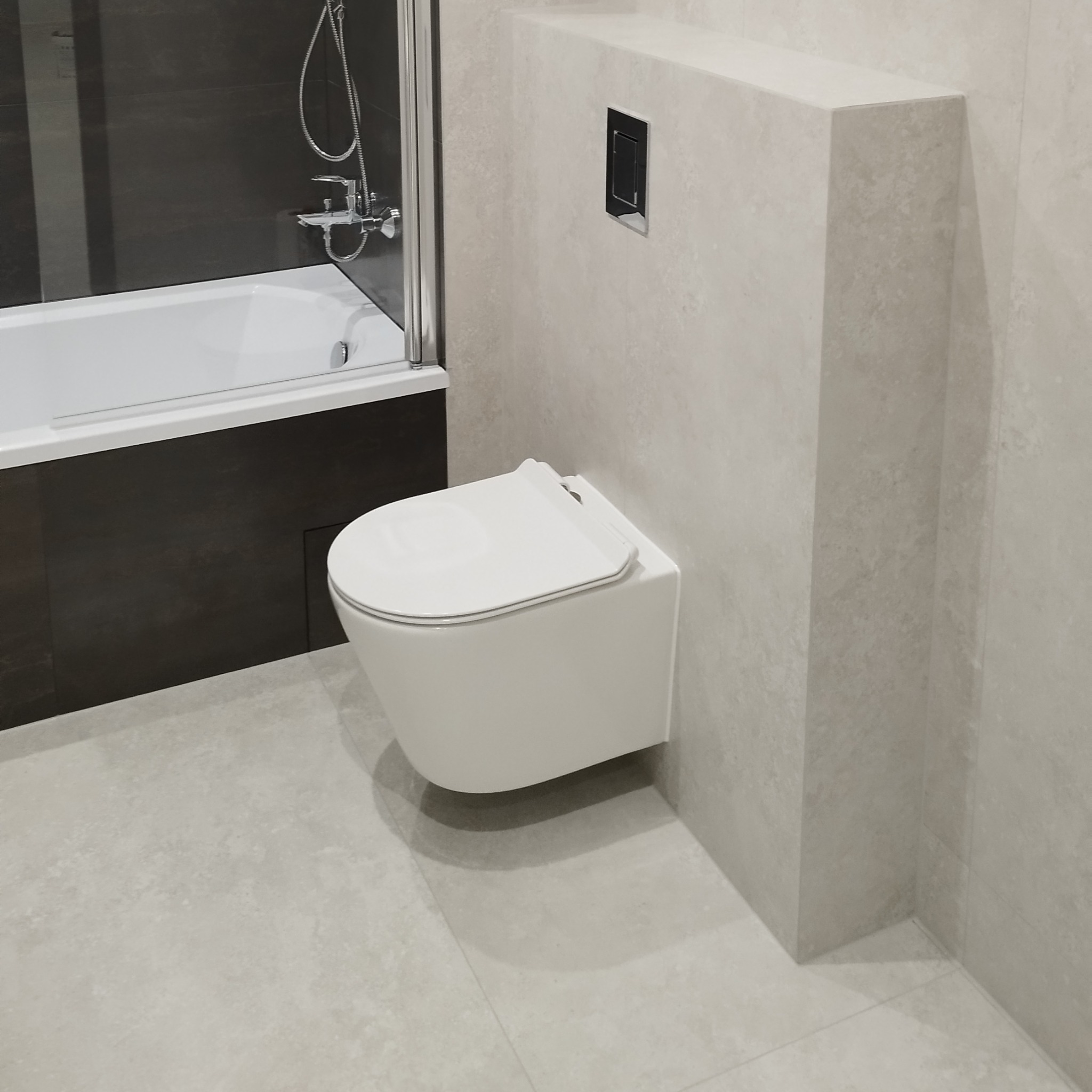 Nowoczesna łazienka z podwieszaną toaletą, zabudowaną spłuczką i płytkami w odcieniach szarości. Widoczna wanna z parawanem nawannowym i chromowaną armaturą.