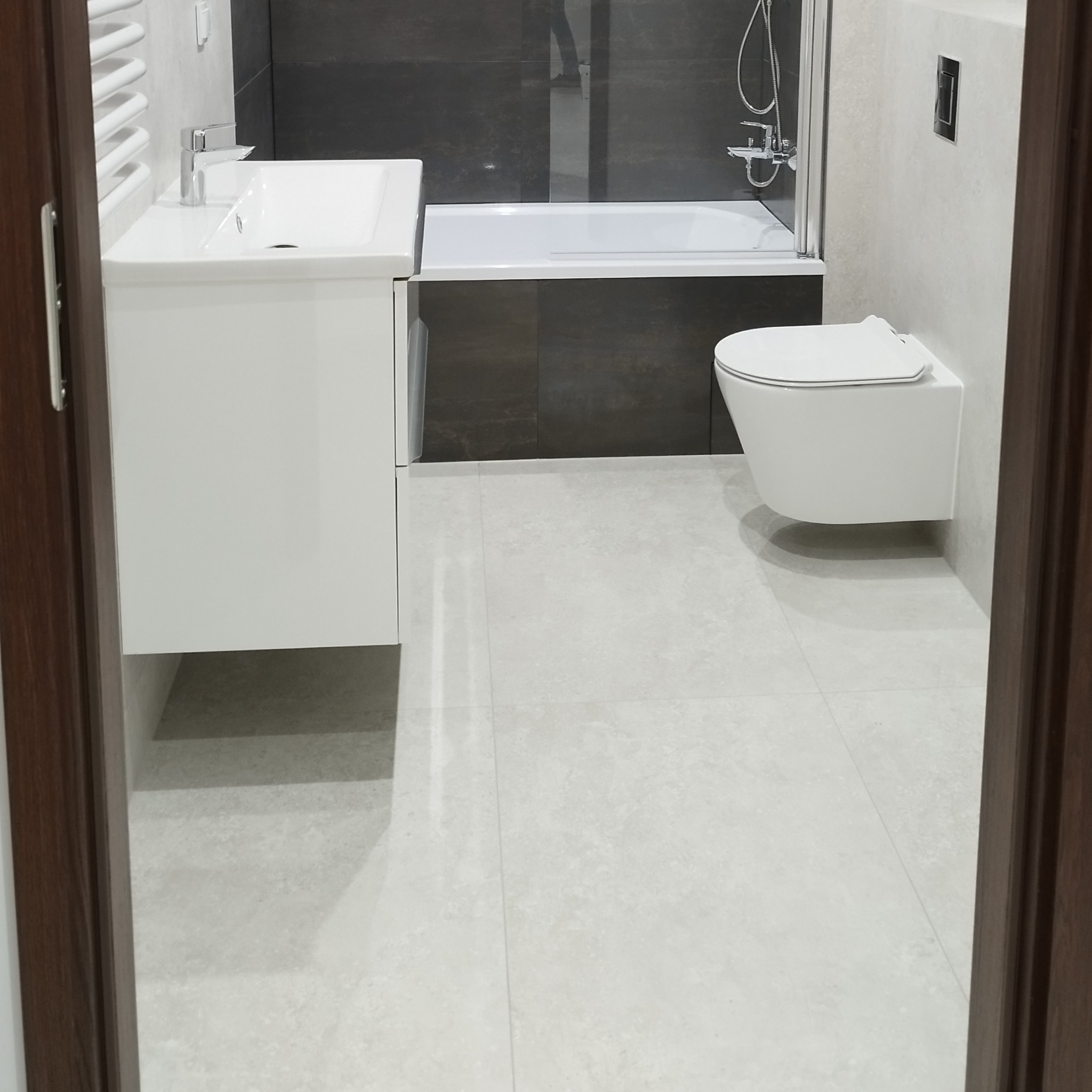 Wykończona łazienka w jasnych odcieniach szarości z podwieszaną toaletą, białą szafką z umywalką i częściowo widoczną wanną z parawanem.