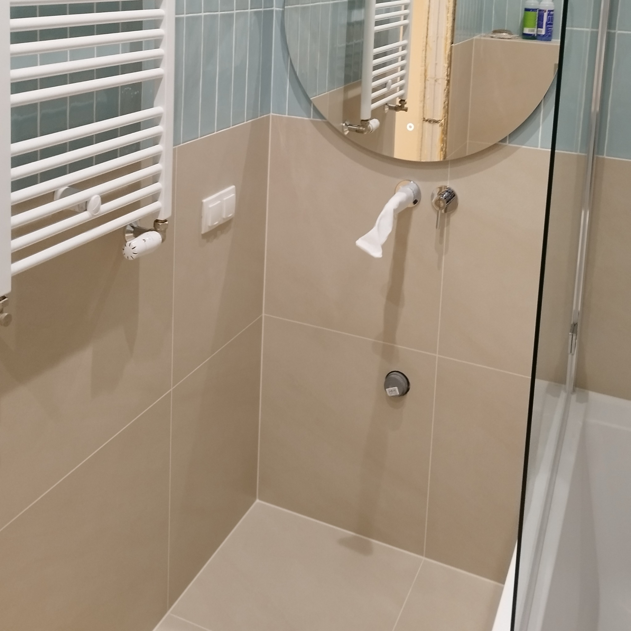 Wykończona łazienka z beżowymi i błękitnymi płytkami, grzejnikiem drabinkowym i okrągłym lustrem z podświetleniem LED. Widoczna armatura i fragment kabiny prysznicowej.