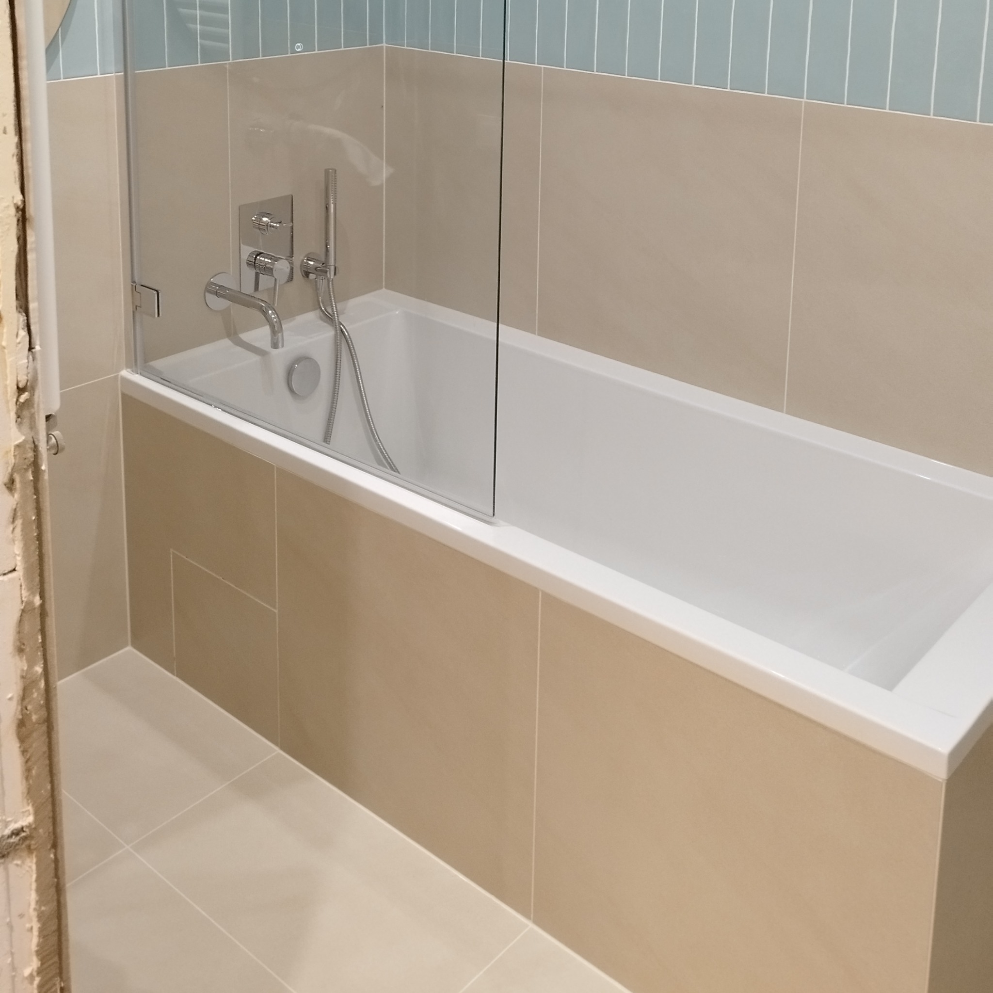 Wykończona łazienka z beżowymi płytkami, białą wanną i szklanym parawanem. Uwagę zwraca niedokończone wykończenie futryny po lewej stronie kadru.