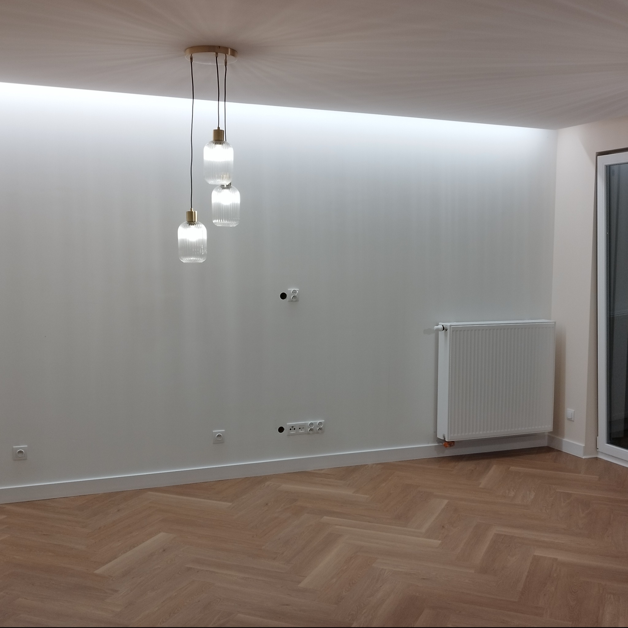 Jasne wnętrze po remoncie: parkiet w jodełkę, białe ściany z gładzią, nowoczesne oświetlenie sufitowe i grzejnik. Widoczne gniazdka elektryczne i listwy przypodłogowe.