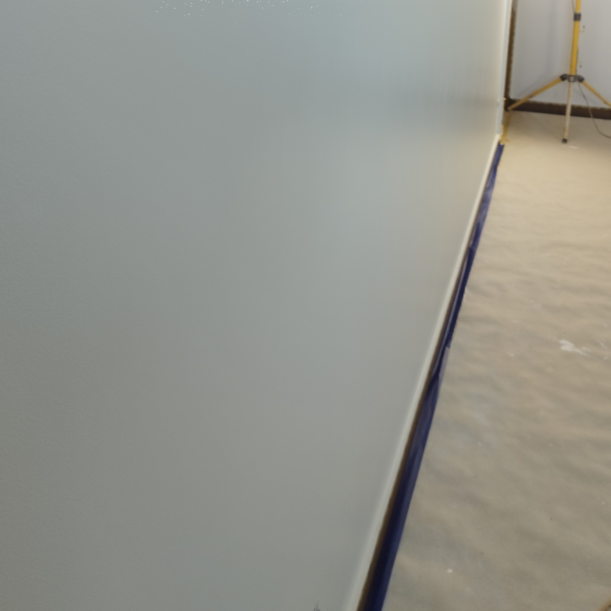 Gładka, świeżo pomalowana ściana w jasnym odcieniu szarości, zabezpieczona niebieską taśmą malarską przy podłodze. W tle widoczny statyw oświetleniowy.