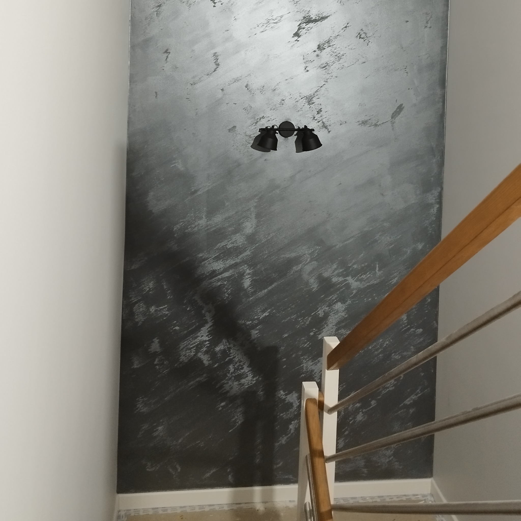 Ściana z efektem dekoracyjnym w odcieniach szarości, oświetlona przez czarny kinkiet. Widoczne fragmenty schodów z drewnianą barierką i metalowymi prętami.