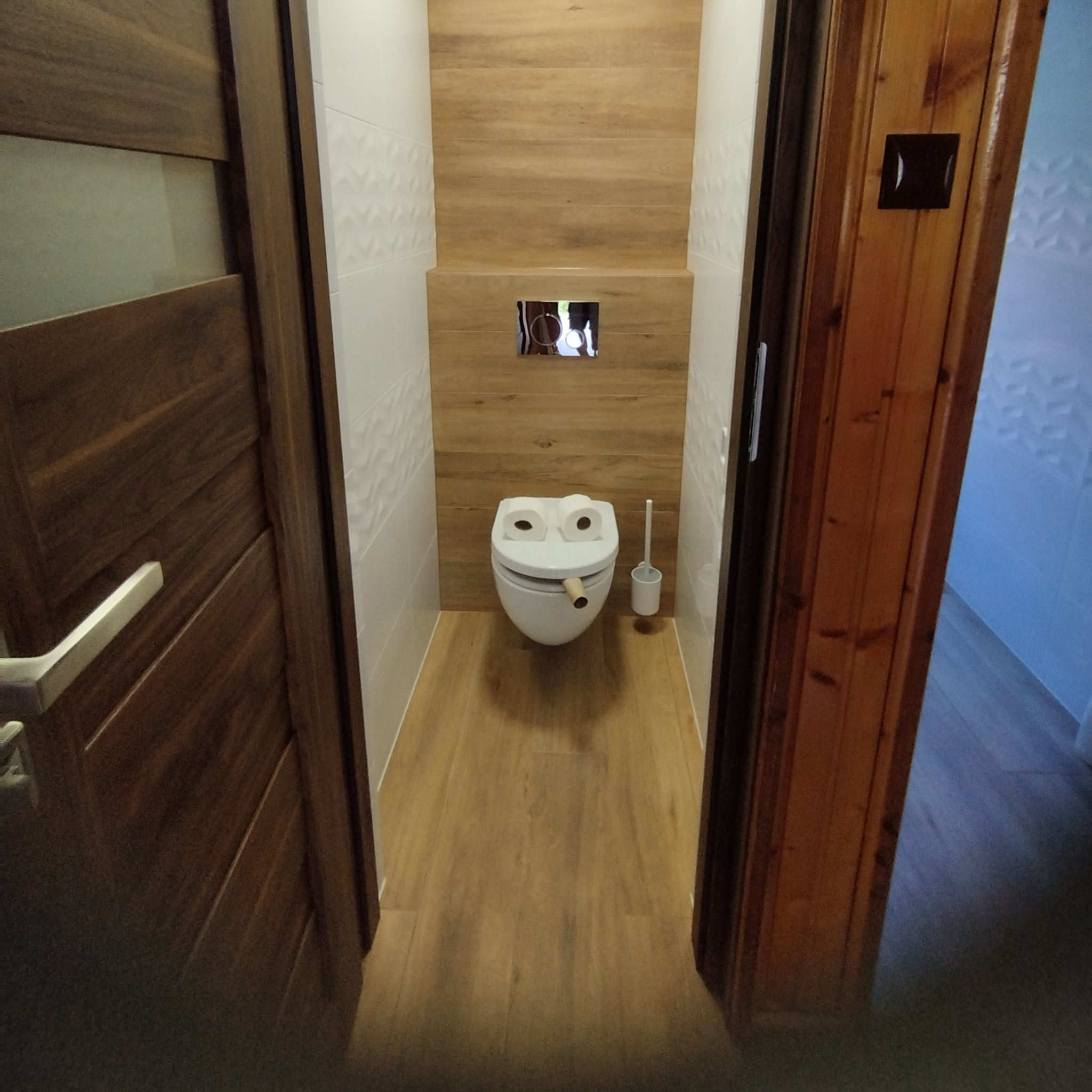 Wąska łazienka z podwieszaną toaletą i drewnianymi akcentami. Białe i drewnopodobne płytki ścienne, drewniana podłoga i drzwi w odcieniach brązu.