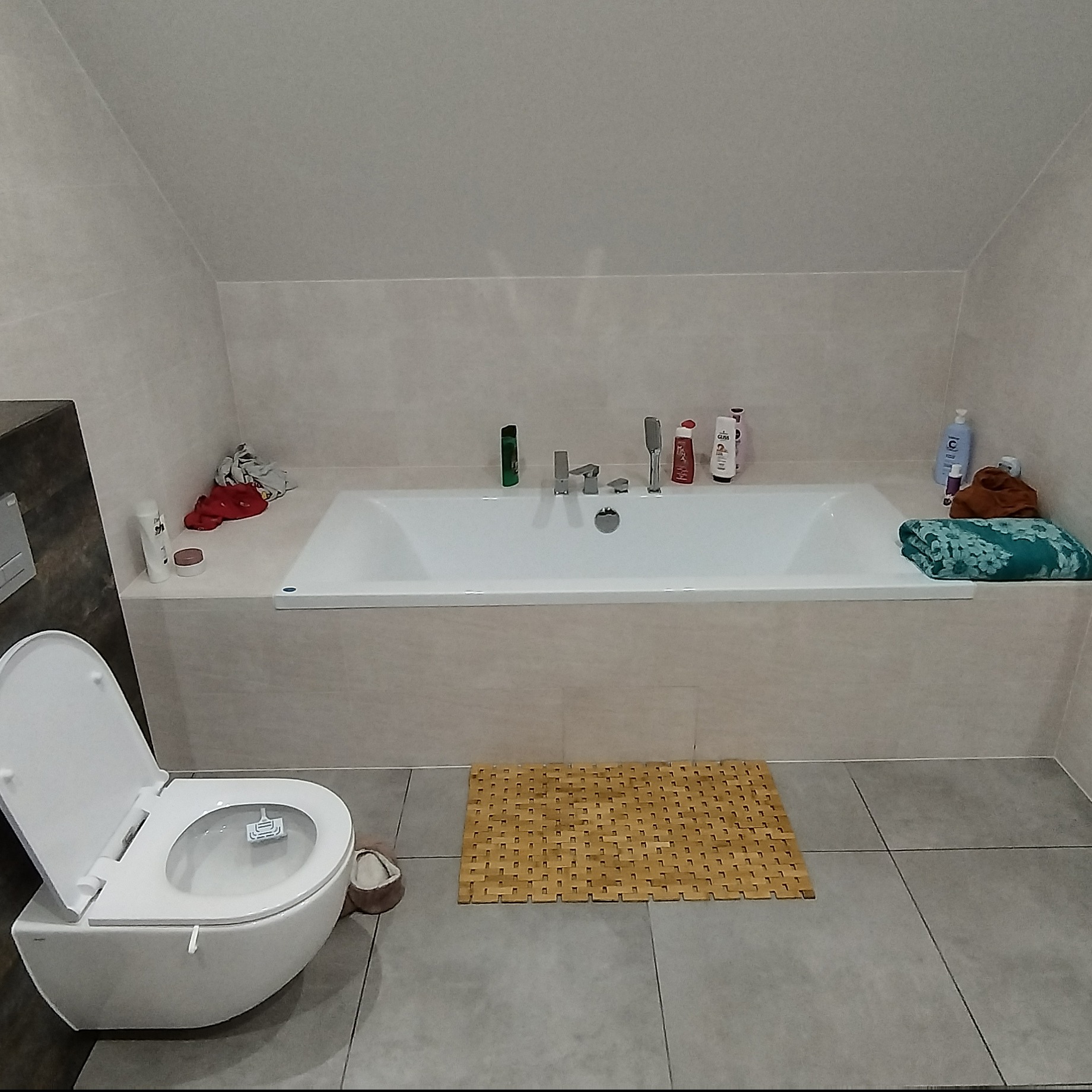 Jasna łazienka na poddaszu z wanną obudowaną płytkami, toaletą i bambusowym dywanikiem. Widoczne kosmetyki i ręczniki. Ukończony remont łazienki.