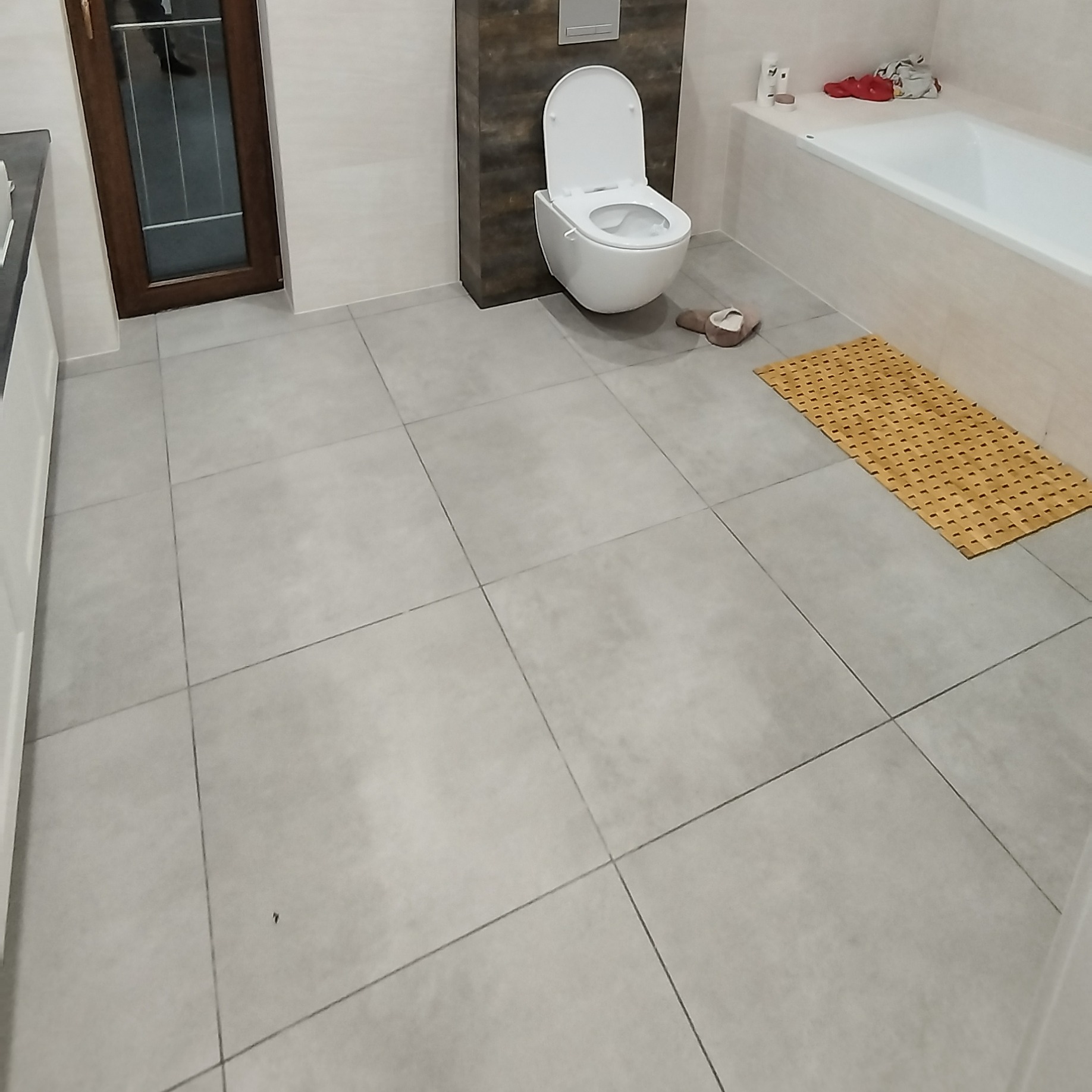 Wykończona łazienka z szarymi, kwadratowymi płytkami podłogowymi, wiszącą toaletą i drewnianą ramą okienną. Minimalistyczny design z akcentem w postaci bambusowej maty.