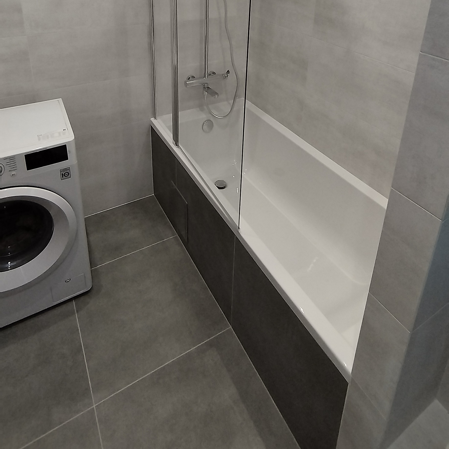 Nowoczesna łazienka w szarościach: biała wanna z szklanym parawanem, ciemnoszara obudowa wanny, pralka LG i szare płytki podłogowe oraz ścienne.