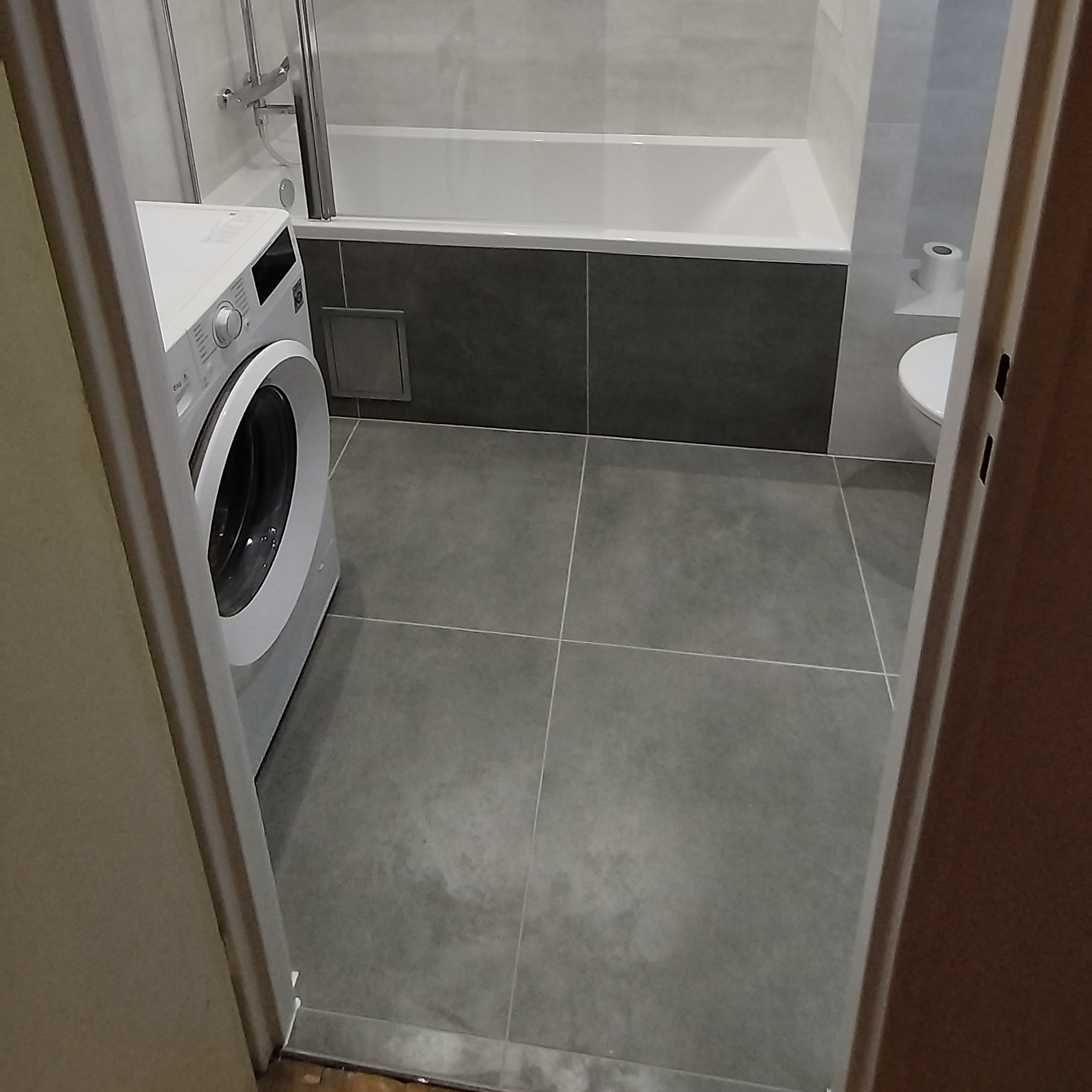 Nowoczesna łazienka z szarymi płytkami, białą wanną i pralką. Widok zza futryny, neutralne oświetlenie. Minimalistyczny design.