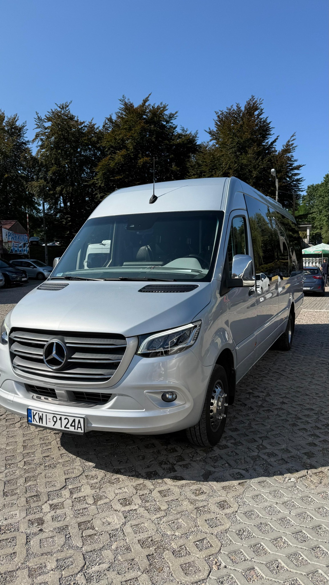 Srebrny bus Mercedes-Benz Sprinter z polską tablicą rejestracyjną na parkingu, widok z przodu, błękitne niebo i drzewa w tle, słoneczny dzień.