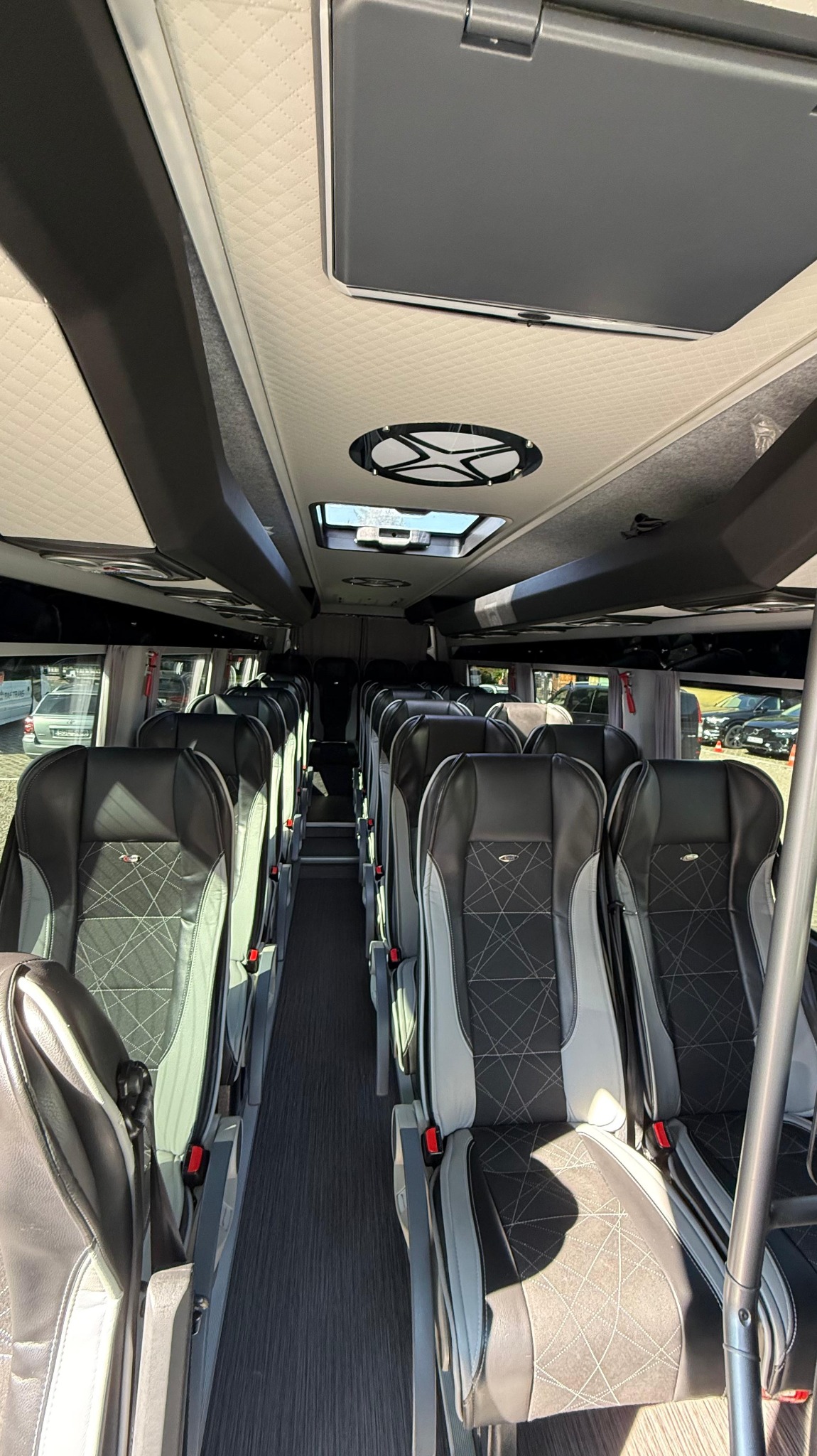Wnętrze luksusowego autobusu turystycznego z rzędami skórzanych foteli w kolorach szarości i czerni, widok z przodu pojazdu, oświetlenie sufitowe i okna dachowe.